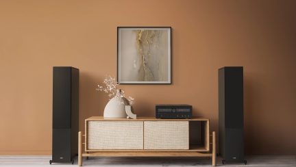 Jasne drewniane meble z plecionymi drzwiami, beżowy wazon z gałązkami, czarny wzmacniacz, 2 czarne głośniki, oprawiony abstrakcyjny obraz