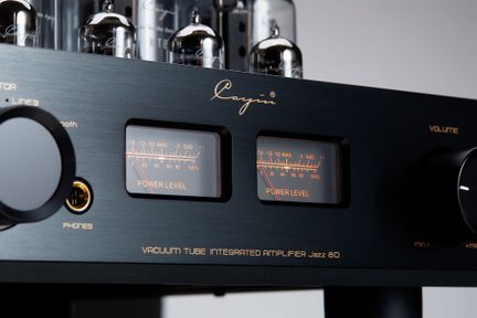 Tube audio amplifier with brushed black metal casing, shown from a three-quarter front angle, featuring two orange backlit VU meters, a glossy black volume knob, a gold headphone jack, and exposed electronic tubes