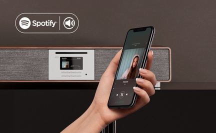 Vue de trois-quarts avant sur une main tenant un smartphone noir en verre et métal affichant un lecteur audio, devant une chaîne hi-fi rectangulaire en métal argenté, bois brun et tissu gris avec fente CD, petit écran couleur et rangée de boutons