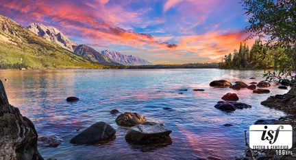 Lac bleu aux reflets rose orangé, bordé de rochers sombres et d’arbres verts, dominé à gauche par une chaîne de montagnes verdâtres et grises sous un ciel crépusculaire multicolore, vue frontale depuis la rive