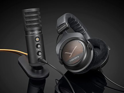 Microfon cilindric negru mat din metal pe suport oval, vedere din trei sferturi, cu grilaj orizontal, indicator galben, două butoane rotative și mufă jack, însoțit de căști circumaurale negre din plastic cu pernițe din catifea, grilaj perforat cuprat inscripționat „TYGR 300 R” și bandă de susținere căptușită, totul așezat pe o suprafață neagră reflectorizantă
