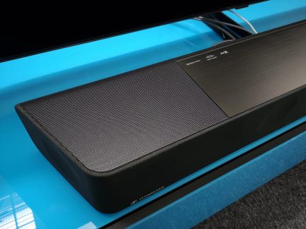 Barra audio nera con griglia metallica sulla parte superiore, posata su una superficie blu lucida