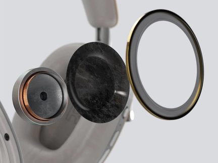 Beige circumaural hörlur i plast och metall, närbild från sidan, exploderad vy av interna komponenter inklusive grå cirkulär driver med kopparkant, svart texturerad dämpningsskiva och transparent ring med guldfärgad metallkant