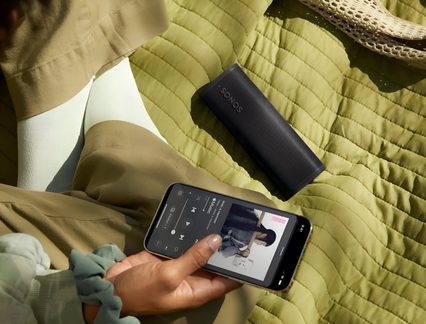 Vue de dessus d’une enceinte cylindrique noire en grille métal perforé avec logo blanc, posée sur un plaid vert matelassé près d’une main tenant un smartphone