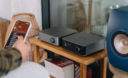 Två grå ljudenheter på en trähylla, en skärm som visar information, en blå högtalare och staplade böcker.