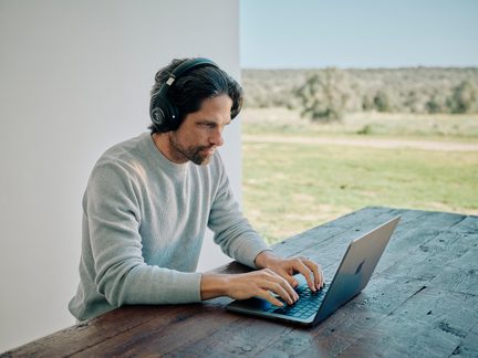 Man med svarta over-ear-hörlurar och ljusgrå tröja, sedd från vänster profil, som skriver på en mörkgrå bärbar dator i aluminium placerad på ett rustikt träbord, med grönskande landskap i bakgrunden