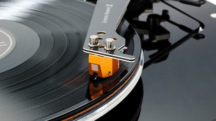 Vue rapprochée en biais d’un bras de lecture métallique noir et argent avec deux vis moletées, équipé d’une cellule orange en contact avec un disque vinyle noir brillant