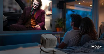 Mały, jasnoszary, sześcienny projektor z tworzywa sztucznego z boczną kratką wentylacyjną, ustawiony na stoliku i widziany z przodu pod kątem, rzucający w tle obraz filmowy przed siedzącą parą.