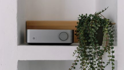 En rektangulär grå enhet med en cirkulär knapp till höger och en hängande grön växt i en kruka till höger på en vit hylla