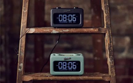 Frontālskats uz diviem taisnstūra digitālajiem radiomodiņiem no plastmasas, no kuriem viens ir matēts melns, bet otrs — matēts salveža zaļš, katrs novietots uz koka kāpņu pakāpiena, ar zilu LED displeju, kas rāda 08:06, un virsū uzstādīti rādītāji.