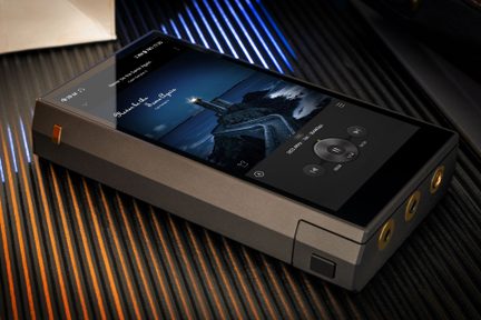 Rectangular portable audio player in anthracite gray metal, illuminated black touchscreen, three gold-rimmed headphone jacks on the front edge, black square button on the side, shown in oblique perspective