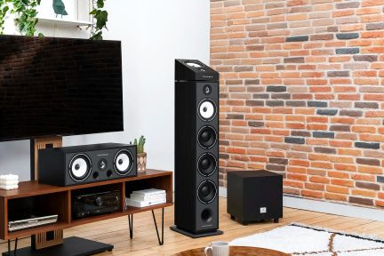 Un téléviseur noir, une enceinte centrale noire, une enceinte colonne noire, un caisson noir, un meuble en bois, mur en briques