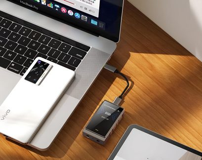 Pogled zgoraj na majhen pravokotni srebrn kovinski avdio napeljnik z črnim zaslonom, na katerem je napisano »DSD«, zlato prednje vtičnico za slušalke in prepleten kabel USB-C, ki napravo povezuje z srebrnim prenosnim računalnikom na svetlo leseni mizi.