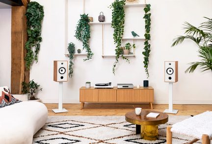 Vista frontale di un soggiorno luminoso con due diffusori rettangolari in legno chiaro con frontale bianco montati su supporti metallici bianchi, un mobile basso in legno chiaro che ospita giradischi e amplificatore, mensole sospese decorate con piante verdi, divano bianco e tavolino rotondo in legno.
