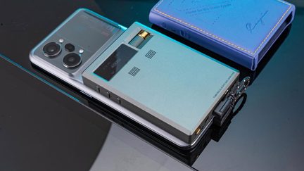 Närbild från ovan av ett silverfärgat glas-smartphone med ett rektangulärt, bärbart ljudförstärkare i ljusblått aluminium ovanpå. Förstärkaren har en liten svart skärm, en förgylld ratt och dubbla galler. Allt ligger på en reflekterande yta bredvid ett blått fodral i konstläder.