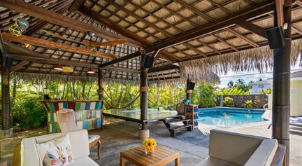 Piscine rectangulaire entourée d'une terrasse couverte en bois avec hamacs, fauteuils, tables basses et végétation tropicale
