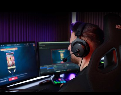 Vue arrière d'une personne portant un casque audio avec micro, assise sur une chaise gaming devant 2 écrans et un panneau lumineux