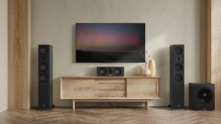 Meuble en bois clair avec 3 tiroirs et 2 portes, téléviseur mural, 2 enceintes noires, caisson de basses, vase beige avec fleurs