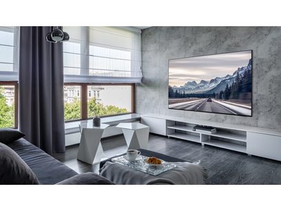 Téléviseur Samsung TQ65S95D 2024 dans un salon moderne avec canapé gris, table basse géométrique et grande baie vitrée donnant sur un paysage montagneux
