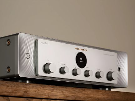 Amplificateur argenté, vue de trois quarts, 6 boutons ronds, écran central noir, logo en haut à droite, posé sur bois