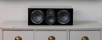 Enceinte noire rectangulaire avec 2 grands haut-parleurs circulaires et 1 tweeter central sur meuble blanc à 3 tiroirs