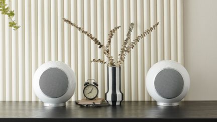Vue de face sur deux enceintes sphériques blanches en plastique brillant avec grille circulaire grise, posées de part et d’autre sur un plateau en bois sombre entourant un vase rayé noir et blanc garni de branches sèches devant un panneau mural rainuré crème