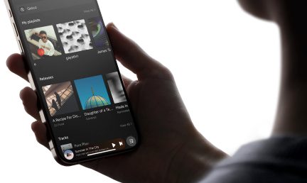 Κύριο άτομο κρατώντας ένα smartphone που εμφανίζει μια μουσική εφαρμογή με λίστες αναπαραγωγής, άλμπουμ και ένα κομμάτι σε αναπαραγωγή ορατό.