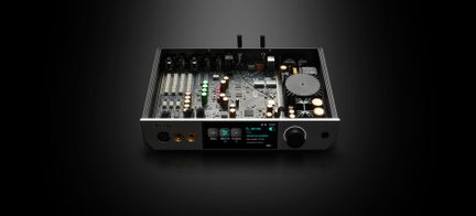 Caja de audio de metal negro y aluminio plateado, vista frontal en ligera vista cenital, tapa retirada que revela la placa electrónica con filas de condensadores dorados y verdes, gran disipador circular y panel frontal equipado con una pantalla a color, un gran selector negro y dos tomas de auriculares doradas.
