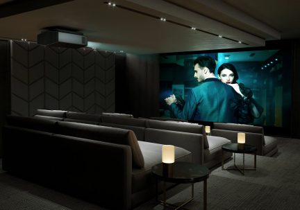 Salle de cinéma privée avec canapés gris, tables rondes noires, lampes cylindriques, écran géant, projecteur au plafond