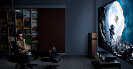 schermo rettangolare molto grande, immagine di fantascienza luminosa, cornice nera, parete grigia, diffusori a colonna in legno e nero, mobile elettronico basso, poltrona design nera e legno, tavolino rotondo, atmosfera scura, vista laterale