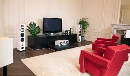Modern living room seen from the front, with a black flat-screen TV on a low black lacquered stand, a pair of white floor-standing speakers with black drivers framing the screen, a white center speaker below, two red cube armchairs on a cream rug, dark wood flooring, and light-colored walls with moldings