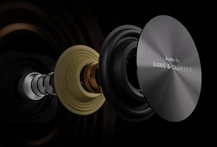 Vue latérale éclatée d’un haut-parleur, succession d’éléments circulaires en métal gris, beige et noir, avec cache avant en aluminium brossé anthracite gravé « Audio by BANG & OLUFSEN »