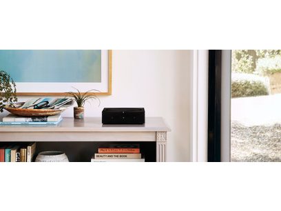 Črn stereo hi-fi ojačevalnik Sonos Amp, postavljen na belo polico, ob knjigah in zeleno lončnico