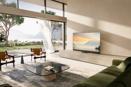 Salon moderne avec grande baie vitrée, 2 fauteuils en bois, table basse en verre, canapé vert, télévision, rideau blanc