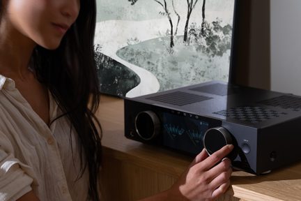 Czarny stereofoniczny wzmacniacz hi‑fi Onkyo na drewnianej szafce, kobieca dłoń regulująca duże pokrętło głośności