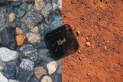 Kamyki i kamienie różnych rozmiarów i kolorów na tle czerwonej ziemi, otwarte czarne pudełko z napisem "Klipsch" położone na wierzchu