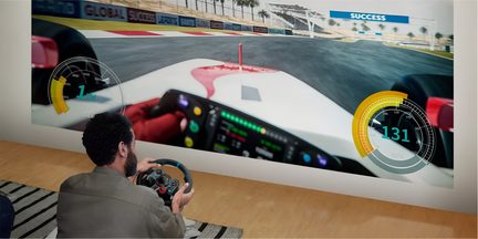 Svart racingsimuleringsratt i plast och metall med flerfärgade knappar, hålls av en man sedd bakifrån framför en stor projicering av ett racingspel
