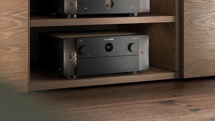 Black Marantz home cinema amplifier with textured aluminum front panel, integrated into a light wood TV cabinet