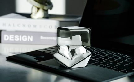 Une paire d'écouteurs blancs dans un boîtier ouvert posé sur un clavier d'ordinateur portable avec des livres en arrière-plan