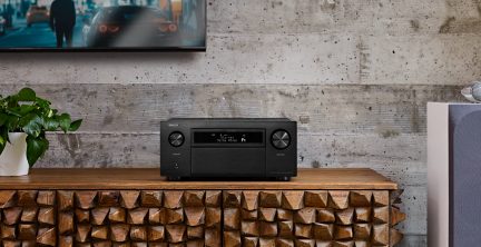 Amplificateur noir avec deux boutons circulaires, écran central, sur meuble en bois sculpté, plante verte en pot blanc à gauche