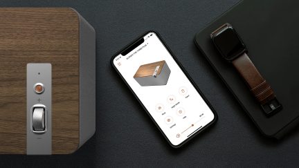 Altavoz rectangular gris, smartphone negro con interfaz de control en pantalla, reloj inteligente marrón y negro colocados sobre una superficie negra