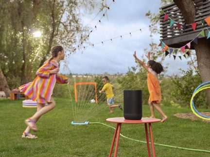 Enceinte cylindrique noire en plastique mat avec grille frontale perforée, vue de face, posée sur une table ronde métallique rouge dans un jardin