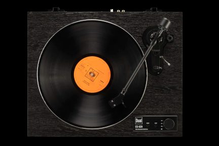Tourne-disque noir finition bois sombre, plateau supportant un disque vinyle noir à étiquette orange, bras de lecture métallique incliné et panneau de commande noir affichant le logo Dual en bas à droite, vue de dessus
