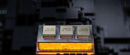 Trīs metāliski pelēkas taisnstūra elektroniskas detaļas ar uzrakstiem, izvietotas uz oranžas caurspīdīgas pamatnes ar apakšas apgaismojumu virs tumša drukas plāksnītes