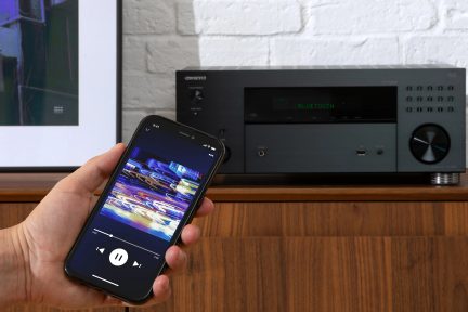 Main view: smartphone displaying music playback screen, black audio receiver with volume knob, white brick wall