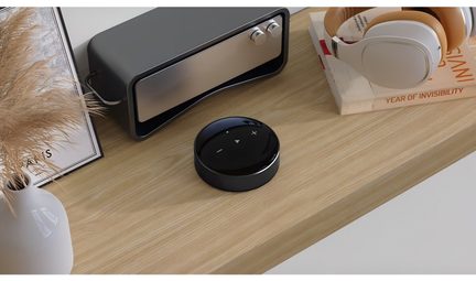 Un disque noir avec des boutons, un appareil rectangulaire gris, un casque blanc, un livre et un vase sur une surface en bois