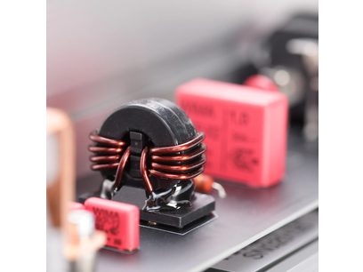 Vedere în plan apropiat oblic a multiprisei de protecție împotriva supratensiunilor Inakustik AC-3200 Filter Analog | SCHUKO, cu bobină toroidală neagră din ferită înfășurată cu fire de cupru emailate, flancată de condensatori dreptunghiulari roz