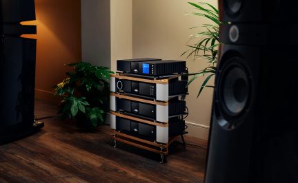 empilage quatre modules hi-fi, faces avant noires, écran central bleu, molettes circulaires argent, châssis cannelés gris foncé, montants latéraux argent, plateaux bois clair, pieds pointus chromés, vue légèrement latérale