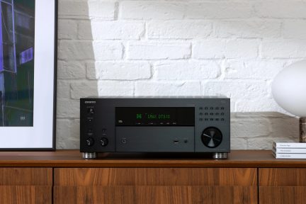 Black amplifier with circular knobs, green screen, on wooden furniture, white brick wall, white lamp, books