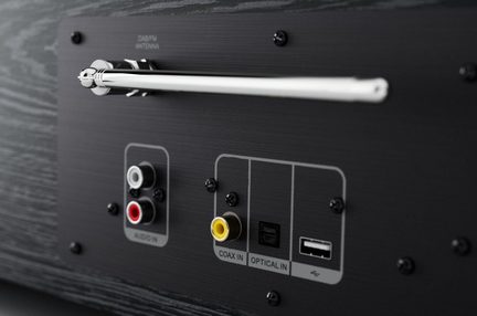 Gros plan, vue de trois-quarts, sur panneau arrière en métal brossé noir d’un boîtier audio à finition bois foncé, avec antenne télescopique argentée horizontale, deux prises RCA rouge et blanche, prise coaxiale jaune, port optique carré noir et port USB rectangulaire blanc